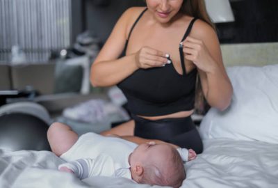 Close-up shot of a mixed race mother unhooking the clasp on her breastfeeding black singlet bra while watching her newborn baby girl lying on the bed.